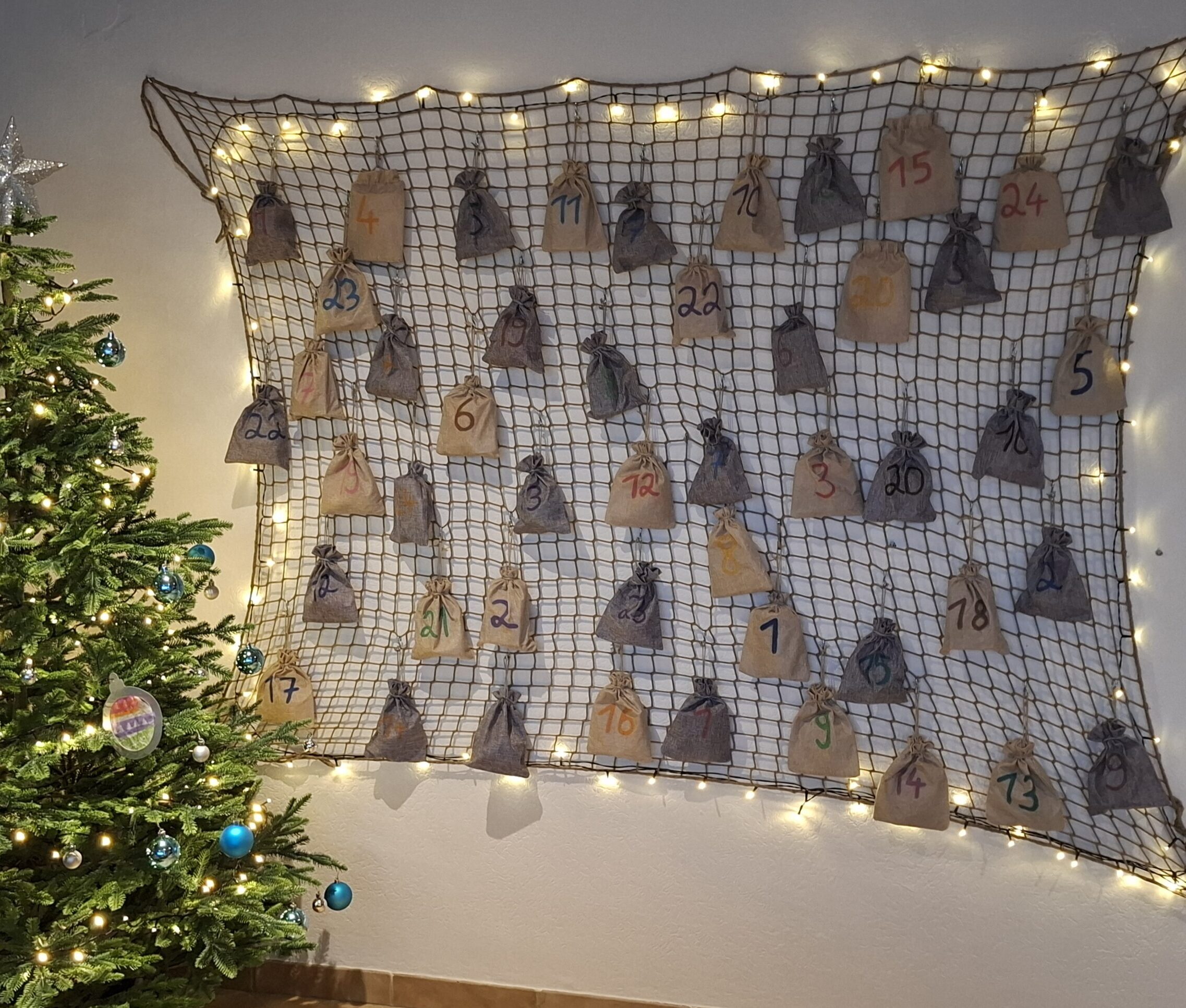 Selbstgemachter Adventskalender aus einem braunen Netz mit beigen und grauen Jutesäckchen und Lichterkette an der Wohnzimmerwand neben einem Tannenbaum
